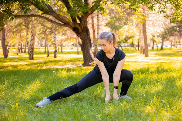 A young, slender woman in a sports uniform does a side lunge exercise to train the muscles of the legs. Fitness classes in nature.  The concept of independent sport training in park.