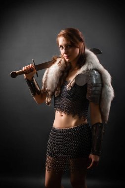 Ortaçağ kadın savaşçısının portresi, zırhlı, omuzlarında kutup tilkisi kürkü elinde bir kılıçla karanlık bir arka planda duruyor. Dişi savaşçı silueti.