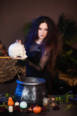 Elinde kafatası olan bir cadının portresi bir kazanın yanında duruyor ve içinde büyücülük için bitkiler ve malzemelerle dolu bir masa..