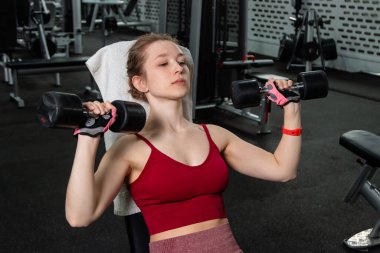 Elinde dambıllar olan genç bir kadın spor salonundaki bankta otururken egzersiz yapıyor..