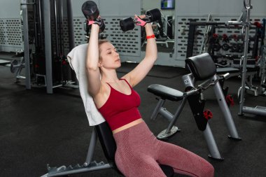 Elinde dambıllar olan genç bir kadın spor salonundaki bankta otururken egzersiz yapıyor..