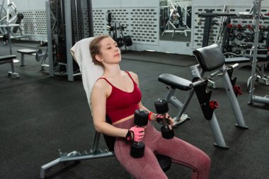 Elinde dambıllar olan genç bir kadın spor salonundaki bankta otururken egzersiz yapıyor..