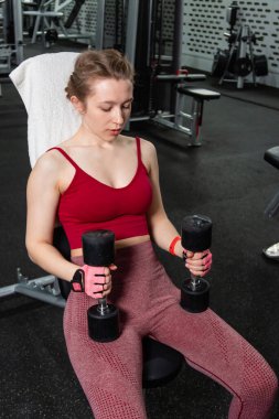 Elinde dambıllar olan genç bir kadın spor salonundaki bankta otururken egzersiz yapıyor..