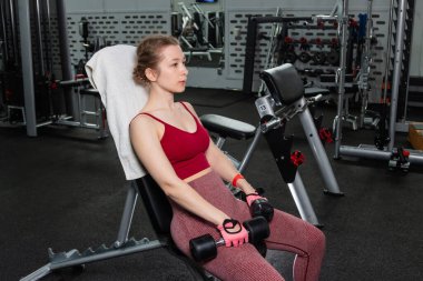 Elinde dambıllar olan genç bir kadın spor salonundaki bankta otururken egzersiz yapıyor..