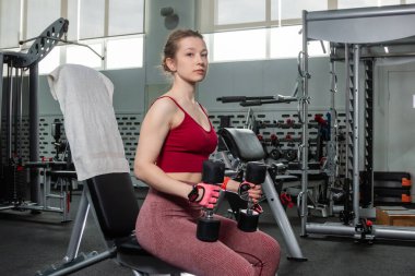 Elinde dambıllar olan genç bir kadın spor salonundaki bankta otururken egzersiz yapıyor..