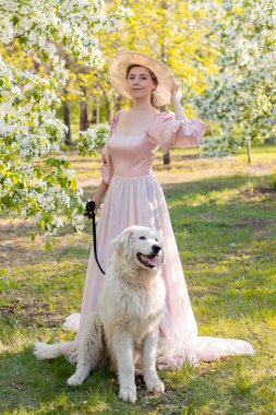 Pembe elbiseli, şapkalı, bahar parkının arka planında duran güzel bir kadının uzun boylu portresi ve tasmalı beyaz köpekli beyaz elma ağaçları.