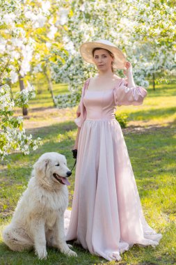 Pembe elbiseli, şapkalı, bahar parkının arka planında duran güzel bir kadının uzun boylu portresi ve tasmalı beyaz köpekli beyaz elma ağaçları.