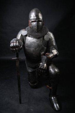 Bir ortaçağ şövalyesinin portresi. Zırhı ve miğferi dolu. Elinde kılıç, diz çökmüş, karanlık bir arka planda izole bir halde. Savaşçı diz çöküyor..