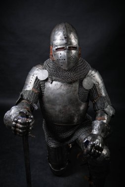 Bir ortaçağ şövalyesinin portresi. Zırhı ve miğferi dolu. Elinde kılıç, diz çökmüş, karanlık bir arka planda izole bir halde. Savaşçı diz çöküyor..