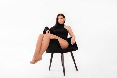 Siyah elbiseli genç bir kadın siyah koltukta beyaz arka planda izole bir şekilde oturuyor..