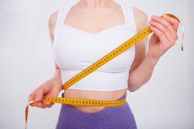 Beyaz arka planda izole edilmiş bir mezura ile belini ölçen güzel bir atletik kadının yakın çekimi. Stüdyoda spor giyim sektöründe çekici bir genç kadın..