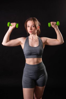 Genç, güzel, atletik bir kız, elinde dambıllarla siyah bir arkaplanda tek başına duruyor. Spor ve sağlıklı bir yaşam tarzı. Bir fitness eğitmeninin portresi.