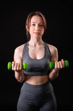 Genç, güzel, atletik bir kız, elinde dambıllarla siyah bir arkaplanda tek başına duruyor. Spor ve sağlıklı bir yaşam tarzı. Bir fitness eğitmeninin portresi.