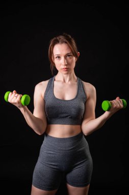 Genç, güzel, atletik bir kız, elinde dambıllarla siyah bir arkaplanda tek başına duruyor. Spor ve sağlıklı bir yaşam tarzı. Bir fitness eğitmeninin portresi.