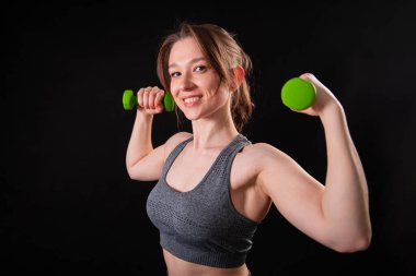 Genç, güzel, atletik bir kız, elinde dambıllarla siyah bir arkaplanda tek başına duruyor. Spor ve sağlıklı bir yaşam tarzı. Bir fitness eğitmeninin portresi.