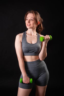 Genç, güzel, atletik bir kız, elinde dambıllarla siyah bir arkaplanda tek başına duruyor. Spor ve sağlıklı bir yaşam tarzı. Bir fitness eğitmeninin portresi.