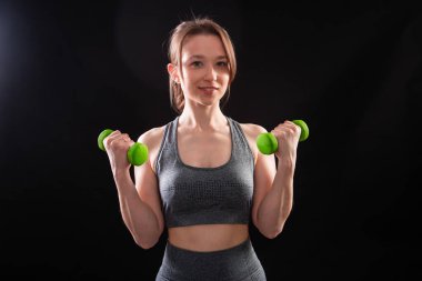 Genç, güzel, atletik bir kız, elinde dambıllarla siyah bir arkaplanda tek başına duruyor. Spor ve sağlıklı bir yaşam tarzı. Bir fitness eğitmeninin portresi.