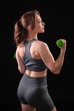 Spor kıyafetli genç bir kadın karanlık arka planda yalnız poz veriyor. Spor salonundaki kız siyah arka planda dambıllarla çalışıyor. Stüdyo çekimi, arka plan, boş alan