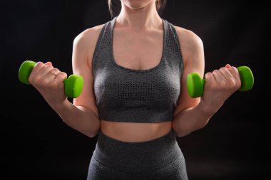 Dambılları tutan bir kadının yakın çekimi. Gri spor giyimli genç bir kadın stüdyoda siyah bir arkaplanda izole bir şekilde poz veriyor. Spor konsepti.