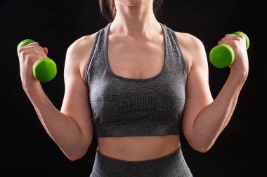 Dambılları tutan bir kadının yakın çekimi. Gri spor giyimli genç bir kadın stüdyoda siyah bir arkaplanda izole bir şekilde poz veriyor. Spor konsepti.