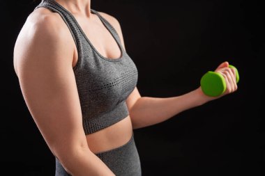 Dambılları tutan bir kadının yakın çekimi. Gri spor giyimli genç bir kadın stüdyoda siyah bir arkaplanda izole bir şekilde poz veriyor. Spor konsepti.