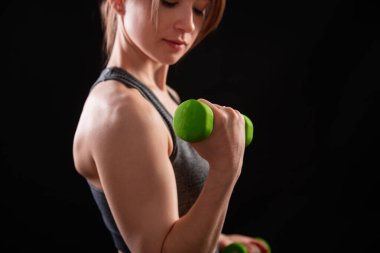 Dambılları tutan bir kadının yakın çekimi. Gri spor giyimli genç bir kadın stüdyoda siyah bir arkaplanda izole bir şekilde poz veriyor. Spor konsepti.