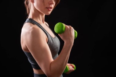 Dambılları tutan bir kadının yakın çekimi. Gri spor giyimli genç bir kadın stüdyoda siyah bir arkaplanda izole bir şekilde poz veriyor. Spor konsepti.