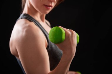 Dambılları tutan bir kadının yakın çekimi. Gri spor giyimli genç bir kadın stüdyoda siyah bir arkaplanda izole bir şekilde poz veriyor. Spor konsepti.