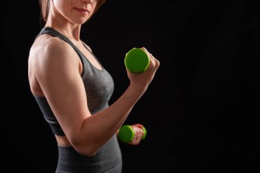 Dambılları tutan bir kadının yakın çekimi. Gri spor giyimli genç bir kadın stüdyoda siyah bir arkaplanda izole bir şekilde poz veriyor. Spor konsepti.