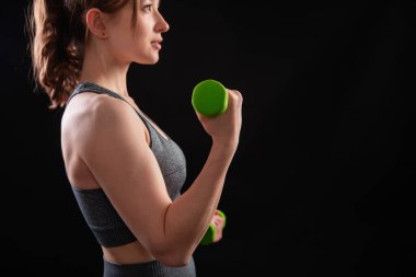 Dambılları tutan bir kadının yakın çekimi. Gri spor giyimli genç bir kadın stüdyoda siyah bir arkaplanda izole bir şekilde poz veriyor. Spor konsepti.
