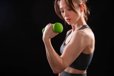 Dambılları tutan bir kadının yakın çekimi. Gri spor giyimli genç bir kadın stüdyoda siyah bir arkaplanda izole bir şekilde poz veriyor. Spor konsepti.