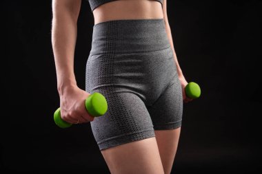 Dambılları tutan bir kadının yakın çekimi. Gri spor giyimli genç bir kadın stüdyoda siyah bir arkaplanda izole bir şekilde poz veriyor. Spor konsepti.