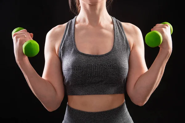 Dambılları tutan bir kadının yakın çekimi. Gri spor giyimli genç bir kadın stüdyoda siyah bir arkaplanda izole bir şekilde poz veriyor. Spor konsepti.