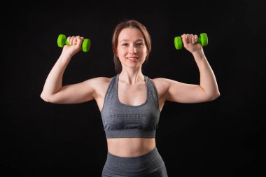 Genç, güzel, atletik bir kız, elinde dambıllarla siyah bir arkaplanda tek başına duruyor. Spor ve sağlıklı bir yaşam tarzı. Bir fitness eğitmeninin portresi.