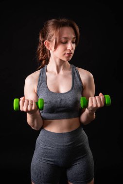 Genç, güzel, atletik bir kız, elinde dambıllarla siyah bir arkaplanda tek başına duruyor. Spor ve sağlıklı bir yaşam tarzı. Bir fitness eğitmeninin portresi.
