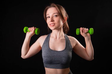 Genç, güzel, atletik bir kız, elinde dambıllarla siyah bir arkaplanda tek başına duruyor. Spor ve sağlıklı bir yaşam tarzı. Bir fitness eğitmeninin portresi.