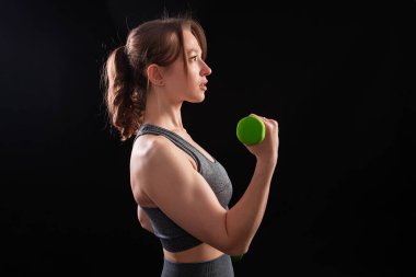 Genç, güzel, atletik bir kız, elinde dambıllarla siyah bir arkaplanda tek başına duruyor. Spor ve sağlıklı bir yaşam tarzı. Bir fitness eğitmeninin portresi.