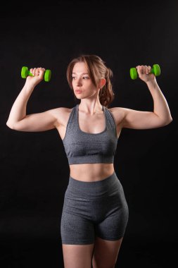 Genç, güzel, atletik bir kız, elinde dambıllarla siyah bir arkaplanda tek başına duruyor. Spor ve sağlıklı bir yaşam tarzı. Bir fitness eğitmeninin portresi.
