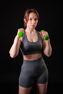 Genç, güzel, atletik bir kız, elinde dambıllarla siyah bir arkaplanda tek başına duruyor. Spor ve sağlıklı bir yaşam tarzı. Bir fitness eğitmeninin portresi.