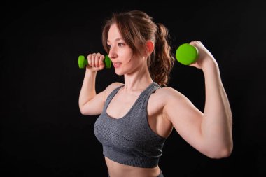 Genç, güzel, atletik bir kız, elinde dambıllarla siyah bir arkaplanda tek başına duruyor. Spor ve sağlıklı bir yaşam tarzı. Bir fitness eğitmeninin portresi.