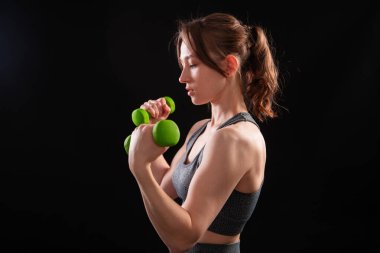 Genç, güzel, atletik bir kız, elinde dambıllarla siyah bir arkaplanda tek başına duruyor. Spor ve sağlıklı bir yaşam tarzı. Bir fitness eğitmeninin portresi.