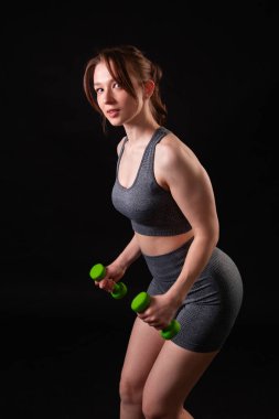 Genç, güzel, atletik bir kız, elinde dambıllarla siyah bir arkaplanda tek başına duruyor. Spor ve sağlıklı bir yaşam tarzı. Bir fitness eğitmeninin portresi.