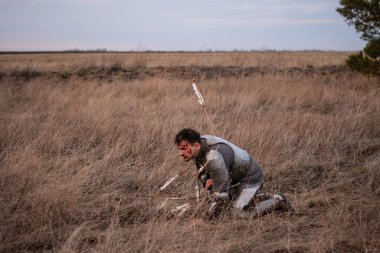Oklarla yaralanmış bir şövalye savaştan sonra kuru çimlerin üzerinde oturur. Yenilmiş bir ortaçağ savaşçısı kanlı yüzüyle bir tarlada diz çöker..