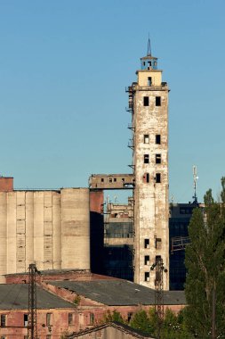 Mavi gökyüzüne karşı eski bir asansör. Ağaçlar ön planda yetişir. Terk edilmiş binalar. Arka planda eski sanayi binaları var. Dikey görünüm