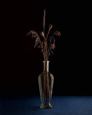 Cam vazoda kurumuş bitkiler. Vazo mavi bir masa örtüsünün üzerinde duruyor. Arkaplan karardı