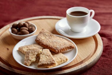 Tabakta kurabiyeler var. Tabağın arkasında küçük bir tabak şeker ve bir fincan kahve var. Tabaklar tahtada. Ahşap bir tahta kırmızı bir masa örtüsünün üzerinde yatıyor..