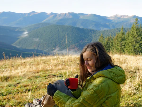 Portrait of girl in yellow down jacket sitting with red cup of hot beverage in the autumn mountains with mist in the valleys