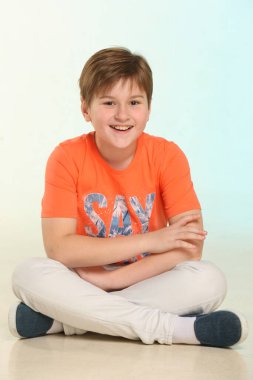 Yakışıklı, modaya uygun, gelişmiş ve turuncu tişörtlü neşeli genç bir çocuk yerde bağdaş kurmuş oturuyor ve neşeyle göz göze gülüyor..