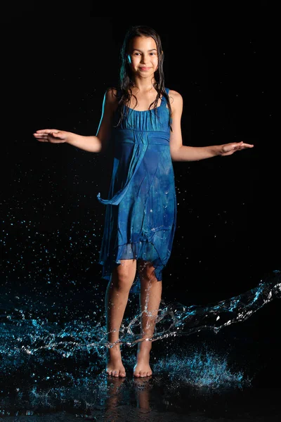 Bright slender beautiful and playful dark-haired teenage girl 12 years old in a blue dress barefoot in a black aqua studio. Emotions in the pouring night rain.