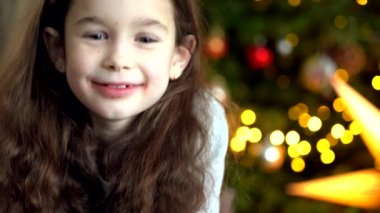 Yeni yıl arifesinde dekore edilmiş odada Noel ağacının yanında oturan mutlu küçük kızın portresi. Çocuk konseptiyle yeni yıl ve tatiller. Bayram kutlamalarının tadını çıkarıyorum. 4k video